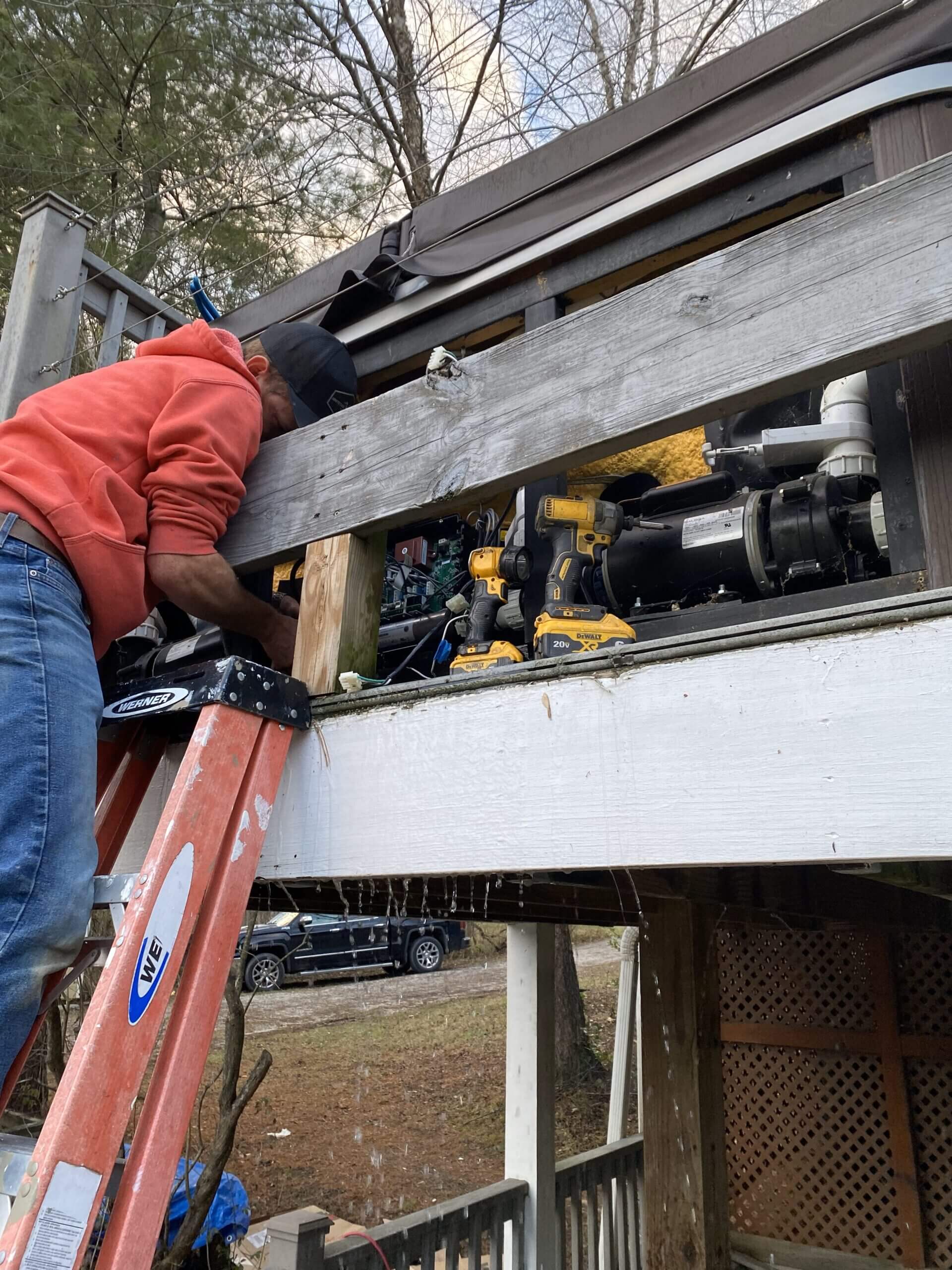 A hot tub repair technician standing on a ladder is repairing a hot tub on a raised deck.